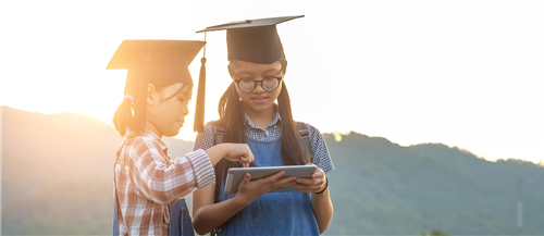 Two young girls wearing graduation caps outside using tablet 